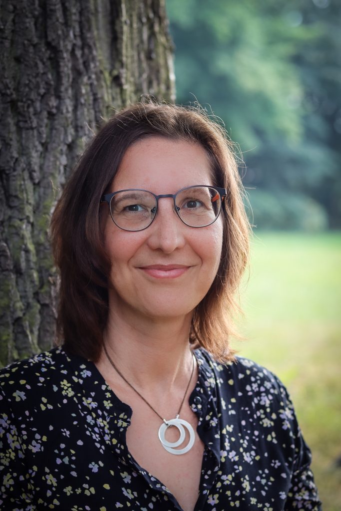 Ein Porträt von Kati Tüngerthal, lächelnd und mit einer Brille, stehend vor einem Baum im Freien. Sie trägt eine dunkel gemusterte Bluse und eine Halskette mit einem doppelten Ringanhänger. Im Hintergrund sind unscharfe grüne Bäume und eine Wiese zu sehen.